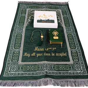 Op de afbeelding: Een donkergroen gebedskleed met een sierlijk patroon, een witte doos met gouden accenten, een groene Koran en een kralen tasbih. De mat heeft tekst in het Engels en Arabisch. De set is voor islamitische religieuze praktijken.