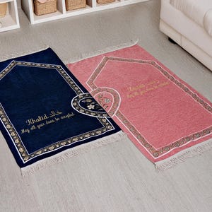 Puede incluir: Dos alfombras de oración, una azul marino y otra rosa, con bordes decorativos y flecos. La alfombra azul marino tiene el nombre "Khalid" y la frase "May all your duas be accepted" en escritura dorada. La alfombra rosa tiene una escritura dorada similar.