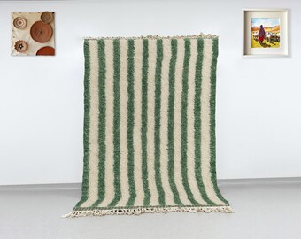 May include: A large, rectangular rug with vertical stripes of green and off-white. The rug has a textured appearance and a fringed edge. Decorative wall art, including framed paintings and woven plates, are visible in the background.