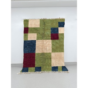 May include: A rectangular rug with a geometric pattern. The rug features squares of green, beige, navy blue, and burgundy. The rug has a shaggy texture and a fringe along the bottom edge. The rug is displayed against a white wall.