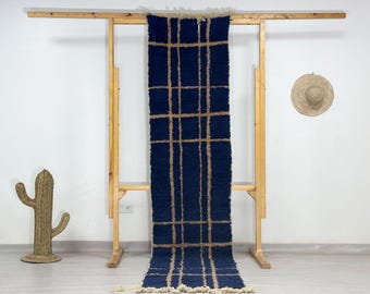 May include: A navy blue and tan checkered rug is displayed on a wooden rack. The rug has a rectangular shape with a grid pattern of tan lines over a dark blue background. The rug is made of a textured material.