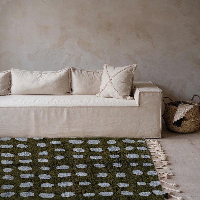 Puede incluir: Escena de sal&oacute;n con un sof&aacute; crema, cojines y una alfombra verde con patrones ovalados blancos. Una cesta de mimbre se coloca a la derecha del sof&aacute;. La alfombra tiene un borde con flecos.