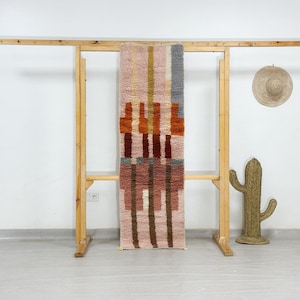 Könnte beinhalten: Ein rechteckiger Teppich mit vertikaler Ausrichtung und geometrischem Muster in Rosa, Orange, Braun und Grau. Der Teppich hängt an einem Holzgestell. Ein gewebter Kaktus und ein Strohhut sind ebenfalls zu sehen.