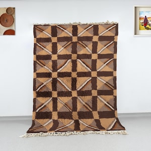 Puede incluir: Una alfombra rectangular grande con un patrón geométrico en tonos marrones y beige. El diseño presenta cuadrados y triángulos, con una franja a lo largo del borde inferior. La alfombra se exhibe contra una pared blanca, con obras de arte a ambos lados.