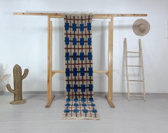 May include: A rectangular rug with a geometric pattern of blue squares and brown lines on a beige background. The rug is displayed on a wooden rack. A woven cactus and a straw hat are also visible.