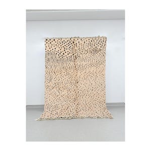 May include: A beige rug with a black and white spotted pattern. The rug has a fringe border.