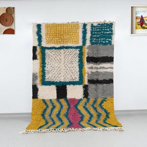 May include: A colorful, geometric rug with a textured, shaggy pile. The rug features a pattern of squares and zigzags in yellow, teal, gray, black, and pink. The rug is displayed against a white wall, with decorative wall art.