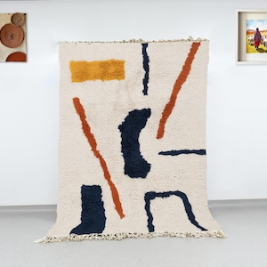 May include: A cream-colored, textured rug with an abstract design. The rug features a mustard-yellow rectangle, navy blue and rust-colored lines. The rug has a fringed edge and is displayed against a white wall.