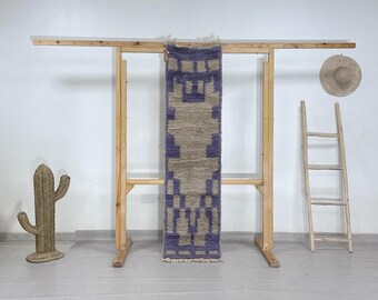 May include: A rectangular, woven rug with a geometric pattern in shades of purple and beige hangs on a wooden rack. A woven cactus and a straw hat are also visible. The rug has a fringed edge.