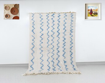 Può includere: Un tappeto color crema con motivi a zig-zag blu. Il tappeto ha un bordo con frange ed è esposto contro un muro bianco. Sono visibili anche opere d'arte murali decorative, tra cui dipinti incorniciati e piatti tessuti.
