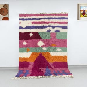 以下が含まれることがあります： 毛足の長いテクスチャのカラフルな幾何学模様のラグ。ピンク、紫、オレンジ、緑など、鮮やかな色の組み合わせが、水平方向と垂直方向のパターンで配置されています。白い壁を背景に展示されています。
