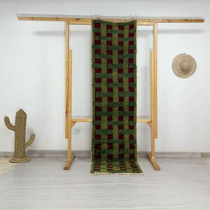 May include: A green and burgundy woven runner rug with a geometric pattern. The rug is displayed on a wooden rack against a white wall. A woven cactus and a straw hat are also visible.