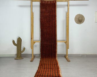May include: A long, rectangular rug with a striped pattern in shades of brown and orange. The rug is displayed on a wooden frame, showcasing its length and texture. The rug has a fringed edge and a neutral background.