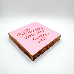 Könnte beinhalten: Ein quadratischer, rosa Holzblock mit dem Text "ZU VIEL NACHDENKEN VERURSACHT GRÜBELKEIT" in Rot. Der Block hat eine hellbraune, unfertige Holzkante.