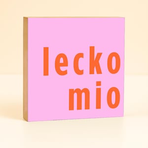 Könnte beinhalten: Ein rosa Holzblock mit den Worten "leckomio" in orangefarbenen Buchstaben. Der Block ist quadratisch und hat einen hellbraunen Rand.