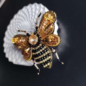 May include: A gold and black beaded bee brooch with a large crystal in the center. The bee's wings are made of gold sequins and the body is made of black beads with gold accents.