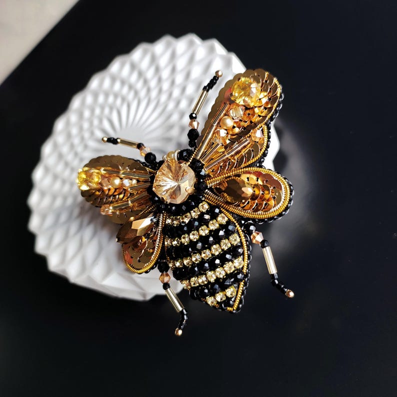May include: A gold and black beaded bee brooch with a crystal body. The bee's wings are made of gold sequins and the body is covered in black beads.
