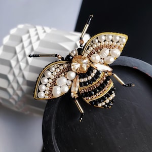 May include: A black and gold bee brooch with a crystal body and pearl accents. The bee is beaded and has a detailed design.