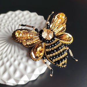 Pode incluir: Um broche de abelha em contas douradas e pretas com um grande cristal no centro. A abelha é detalhada com lantejoulas douradas e contas pretas.