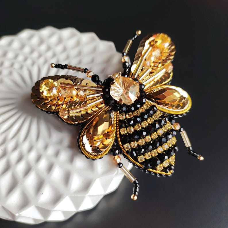 May include: A gold and black beaded bee brooch with a large crystal in the center. The bee is detailed with gold sequins and black beads.