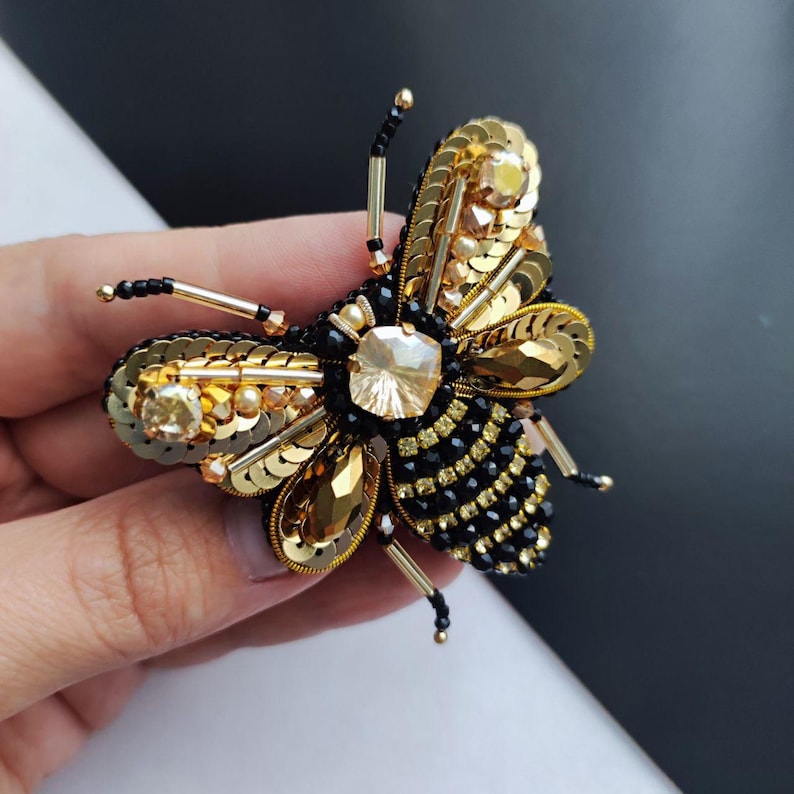 May include: A gold and black beaded bee brooch with a large crystal in the center. The bee has a detailed design with sequins and beads.