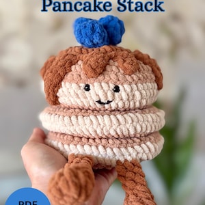 Peut inclure: Peluche empilée de crêpes au crochet avec un visage souriant, des accents bruns de « sirop » et bleus de « myrtille ». Le texte « Beatrix Bluberry Pancake Stack » est affiché en haut et « PDF Pattern » en bas. La peluche est tenue dans une main.