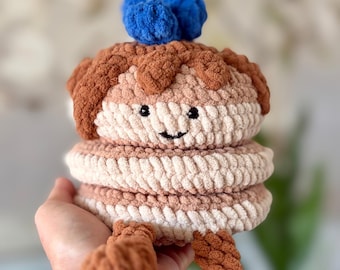 May include: A crocheted pancake stack plushie with a smiling face, brown 'syrup' and blue 'blueberry' accents. The text "Beatrix Bluberry Pancake Stack" is displayed at the top, and "PDF Pattern" is at the bottom. The plushie is held in a hand.