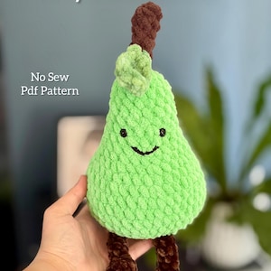 May include: A handmade green crochet pear plushie with a smiling face, brown stem, and brown feet. The text "Juicy Pear Pattern" and "No Sew Pdf Pattern" are visible in the image. The plushie is designed to look like a pear.