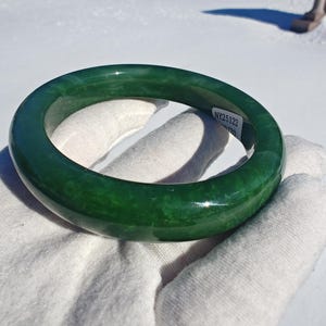 Può includere: Un braccialetto bangle di giada verde vibrante. La pietra lucidata ha una finitura liscia e lucida. Il braccialetto è circolare e sembra essere un pezzo unico di giada. Una piccola etichetta con il testo "NY25122" è visibile.