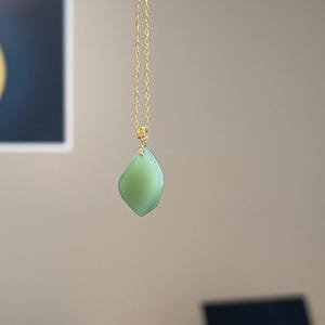 May include: A necklace featuring a smooth, light green, diamond-shaped pendant suspended from a delicate gold-coloured metal chain. The pendant has a polished finish and is attached to the chain by a small, textured gold-coloured metal bail.
