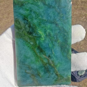 May include: A rectangular jade stone with a polished surface. The stone features a swirling pattern of green and teal hues. The edges are smooth and rounded. The stone is held by a gloved hand.