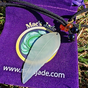 May include: A translucent, pale green jade feather pendant rests on a purple velvet pouch. The pouch features the text "Mac's Jade" and a website address. A black cord with decorative beads and a purple drawstring complete the presentation.