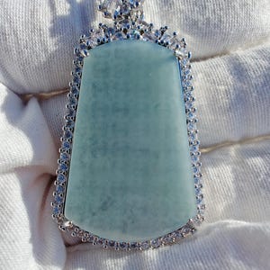 May include: A silver pendant with a large, light green rectangular stone. The stone is surrounded by a border of small, clear crystals. The top of the pendant features a cluster of clear crystals and a small green stone.