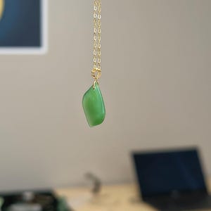 以下が含まれることがあります： ゴールドトーンのチェーンネックレスで、滑らかで不規則な形の緑色のペンダントが一つ付いています。ペンダントは小さなゴールドトーンの丸カンから吊り下げられ、繊細な金属製のチェーンに取り付けられており、フレームの中央に垂直に垂れ下がっています。