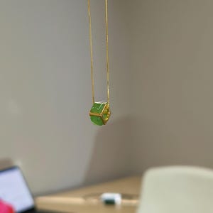 May include: A gold chain necklace featuring a small, hexagonal green jade bead encased in a gold metal frame. The pendant hangs vertically from a thin, delicate gold-toned chain, showcasing a smooth, polished surface on the green stone.