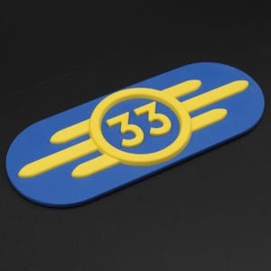 May include: A blue and yellow Vault-Tec emblem with the number 33 in the center. The emblem is a stylized image of a vault door with a glowing yellow symbol.