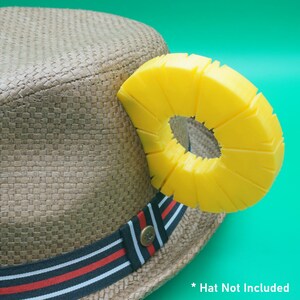 K&ouml;nnte beinhalten: Ein Stroh-Fedora-Hut mit einem schwarz-rot-wei&szlig; gestreiften Band und einem hellgelben Ananas-f&ouml;rmigen Accessoire. Der Text "* Hat Not Included" befindet sich unten rechts.