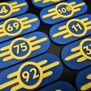 May include: A set of ten blue and yellow Fallout Vault-Tec badges with numbers 4, 69, 75, 92, 108, 11, 3, 5, 7, and 9.