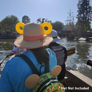 K&ouml;nnte beinhalten: Ein Stroh-Sonnenhut mit einem rosa Band und gelben Mickey Mouse Ohren wird von einer Person auf einem Boot getragen. Der Hut ist der Mittelpunkt, mit dem Text "Hat Not Included" unten. Andere Personen sind im Boot, und ein Schiff ist im Hintergrund.