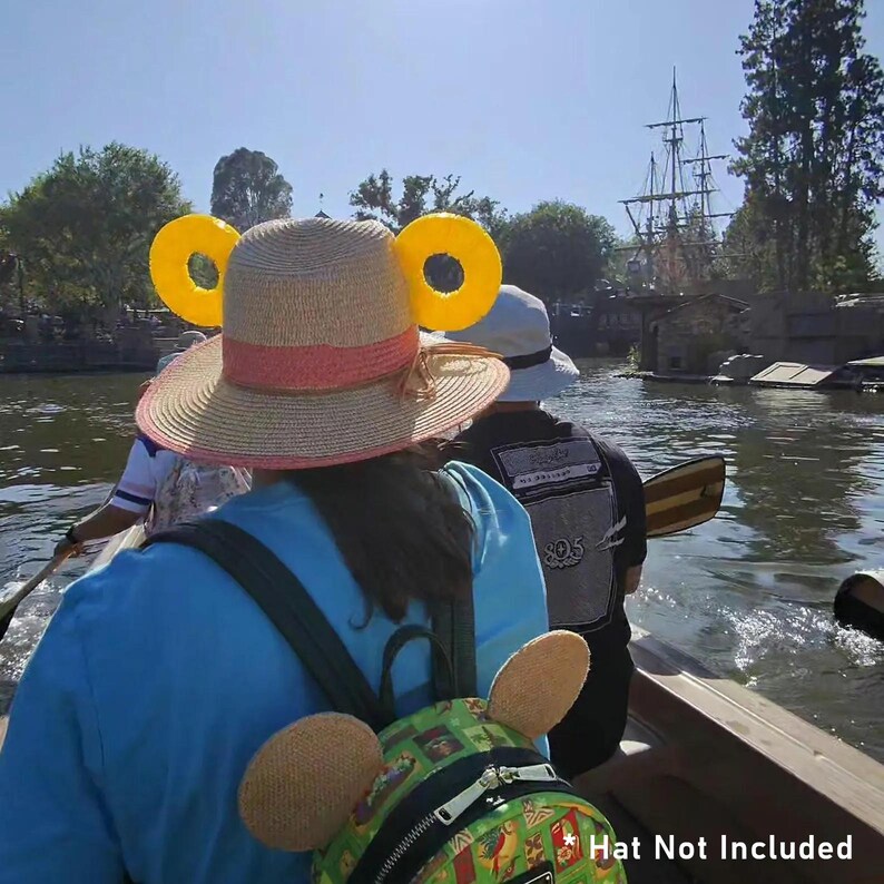 K&ouml;nnte beinhalten: Ein Stroh-Sonnenhut mit einem rosa Band und gelben Mickey Mouse Ohren wird von einer Person auf einem Boot getragen. Der Hut ist der Mittelpunkt, mit dem Text "Hat Not Included" unten. Andere Personen sind im Boot, und ein Schiff ist im Hintergrund.