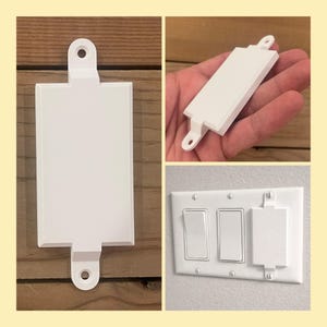 May include: White 3D-printed light switch guard. The guard has a rectangular shape with a small tab and a hole at the top and bottom for mounting. The image shows the guard on a wooden surface, held in a hand, and installed on a light switch plate.