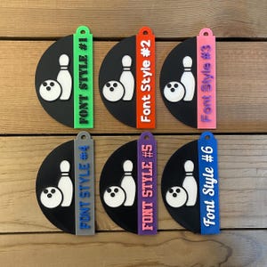 May include: Six bowling-themed keychains. Each keychain features a black semi-circle with a white bowling pin and ball. The keychains have different colored tags with the text "Font Style #" followed by a number.