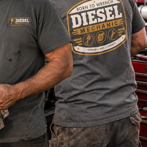 May include: Two gray t-shirts with the text "DIESEL MECHANIC" and related graphics. One shirt has the text on the front, the other on the back. The shirts are worn by people in work pants.