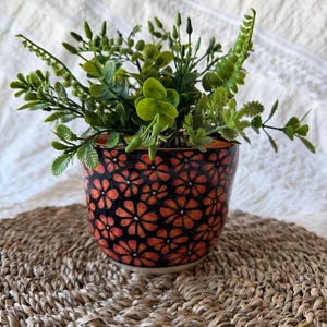 May include: A ceramic flower pot with a black background and orange floral pattern, filled with various green plants. The pot sits on a woven, round placemat. The background is a white, textured surface.