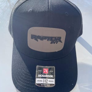 May include: Black mesh trucker hat with a brown patch that says "RAPTOR SVT" in black lettering. The hat has a red and white label that says "EST. 1970 RICHARDSON ORIGINAL 112 TRUCKER ADJUSTABLE".