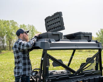 May include: A black utility vehicle with a black roof rack and two black storage boxes. The storage boxes are open and the lid of one is propped up. A person is standing next to the vehicle and is opening the lid of one of the storage boxes.