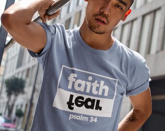 Op de afbeelding: Lichtblauw T-shirt met de tekst "faith teal" in een wit en zwart rechthoekig ontwerp. De tekst "psalm 34" staat eronder. Het T-shirt heeft korte mouwen.