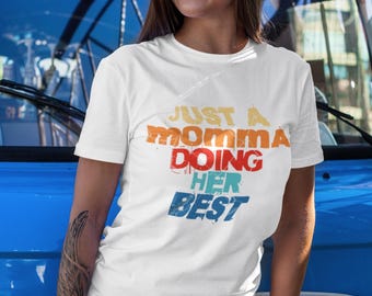 May include: White t-shirt with the text "JUST A MOMMA DOING HER BEST" in orange, red, and blue. The person is wearing sunglasses and denim shorts. A red can is visible.