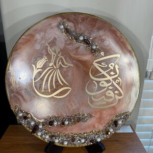 May include: A round decorative piece with a pink and gold resin design. It features gold calligraphy and a dancing figure. Embellished with pearls and crystals, it sits on a black stand, showcasing an artistic and cultural theme.