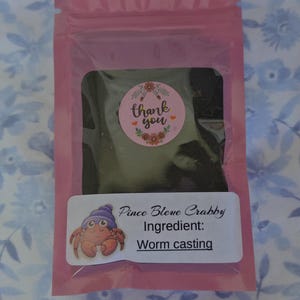 Peut inclure: Un emballage rose contenant une substance sombre, avec un autocollant rond disant "merci" et un motif floral. Une &eacute;tiquette rectangulaire pr&eacute;sente un crabe de dessin anim&eacute; et le texte "Pince Bleve Crabby Ingr&eacute;dient: Worm casting."